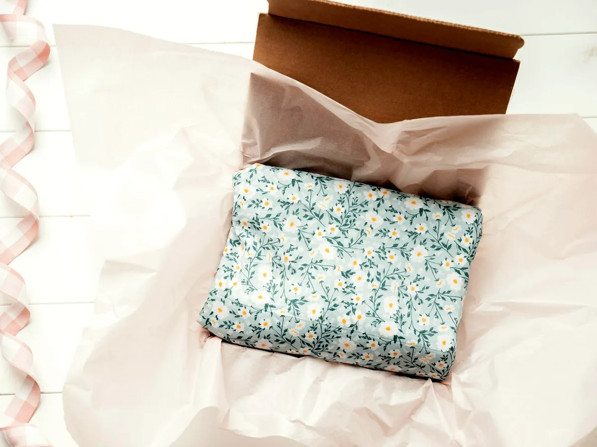 An opened kraft mailer box on a white surface with pink tissue paper, ribbon and a floral-print wrapped item inside.