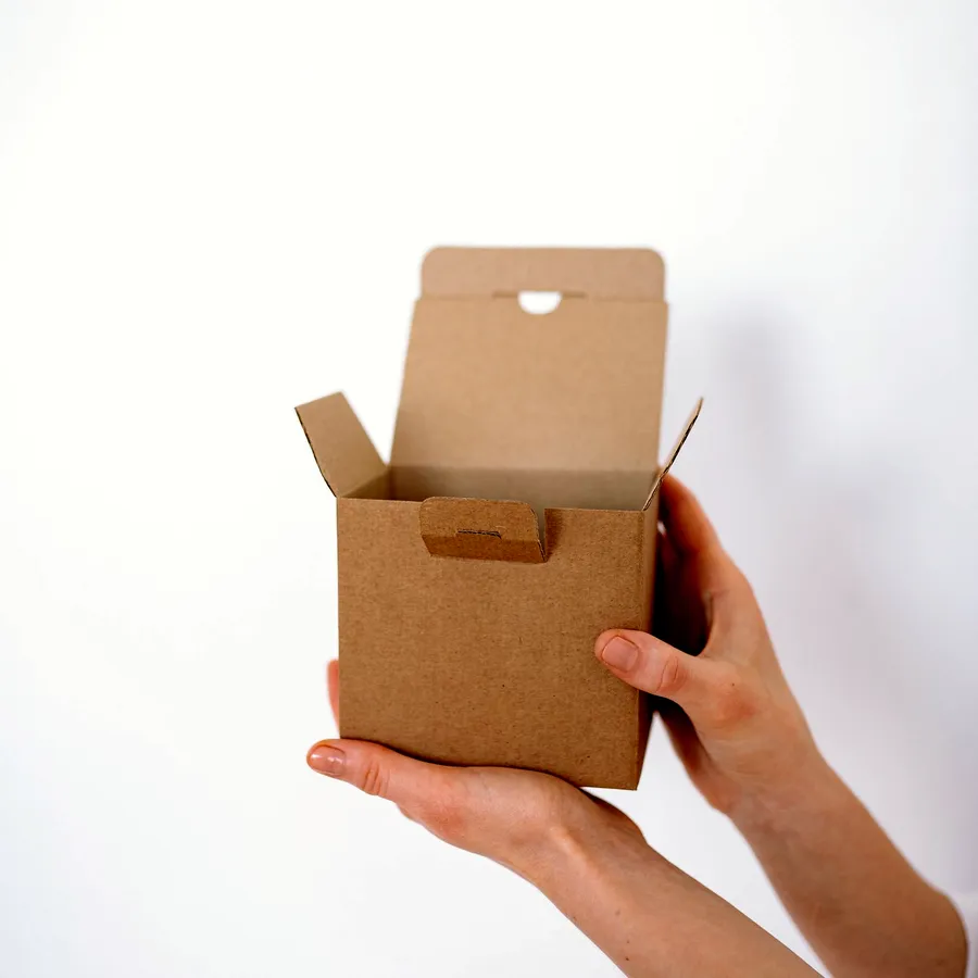 Hands holding a small kraft box with a die-cut tuck flap open against a white wall.